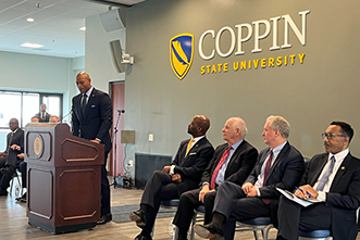 Man on a podium with other men on chairs, COPPIN University sign on the wall