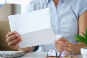 Person holding up a piece of paper reading it with a desk containing a pair of glasses, a laptop and a small plant