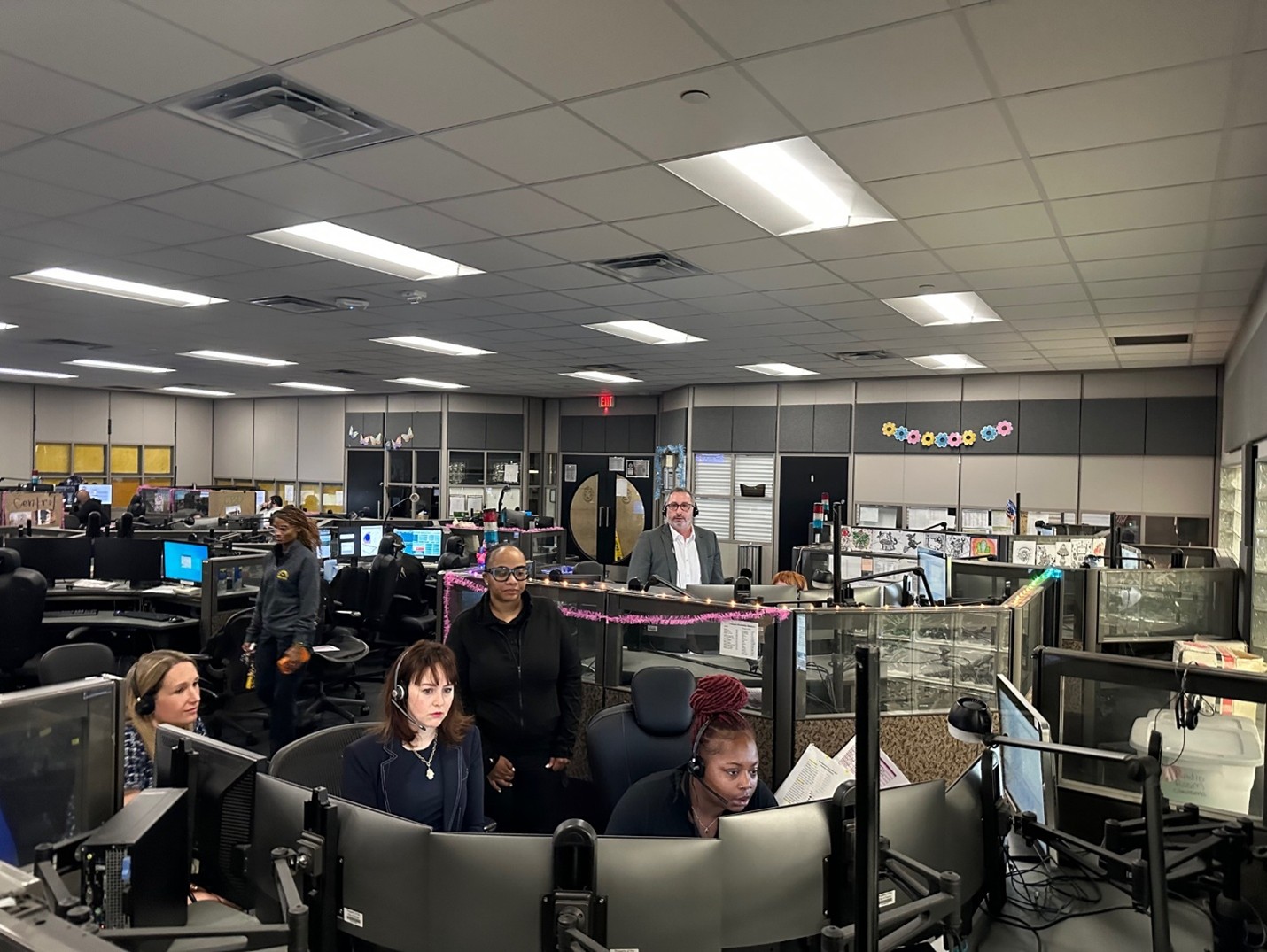 Assistant Secretary Roth and three others are wearing headphones and watching multiple screens inside a 9-1-1 center